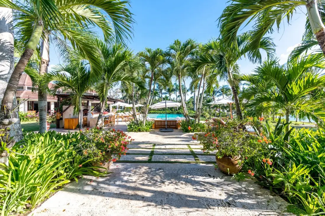 A tropical garden path leads toward a pool, bordered by palms and flowering plants.