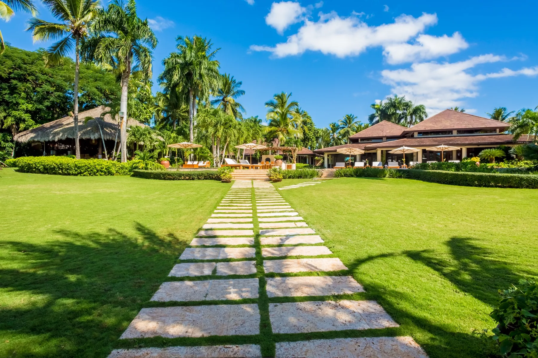 A stone path crosses a lawn toward a tropical resort building surrounded by palm trees.