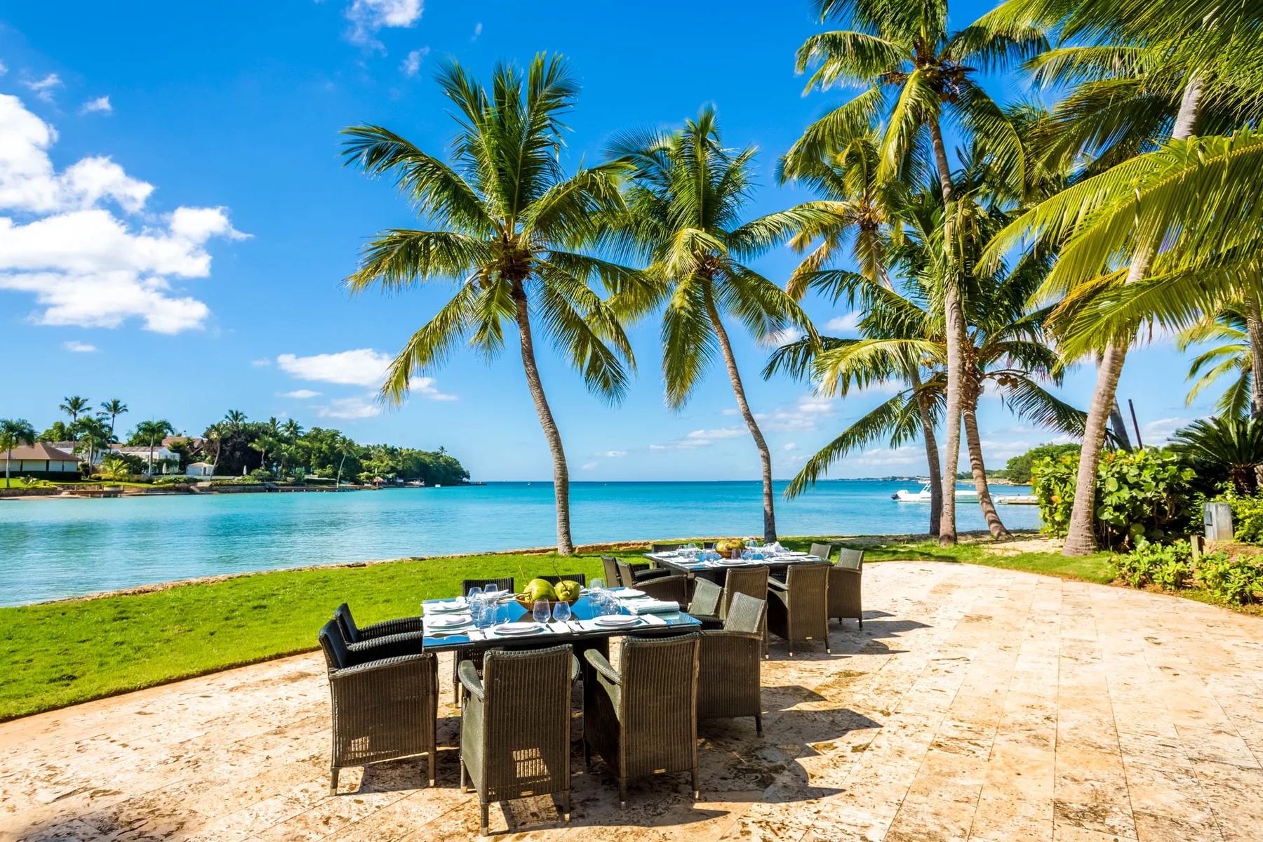 An outdoor dining area beside a tropical shoreline, with palm trees and water beyond.