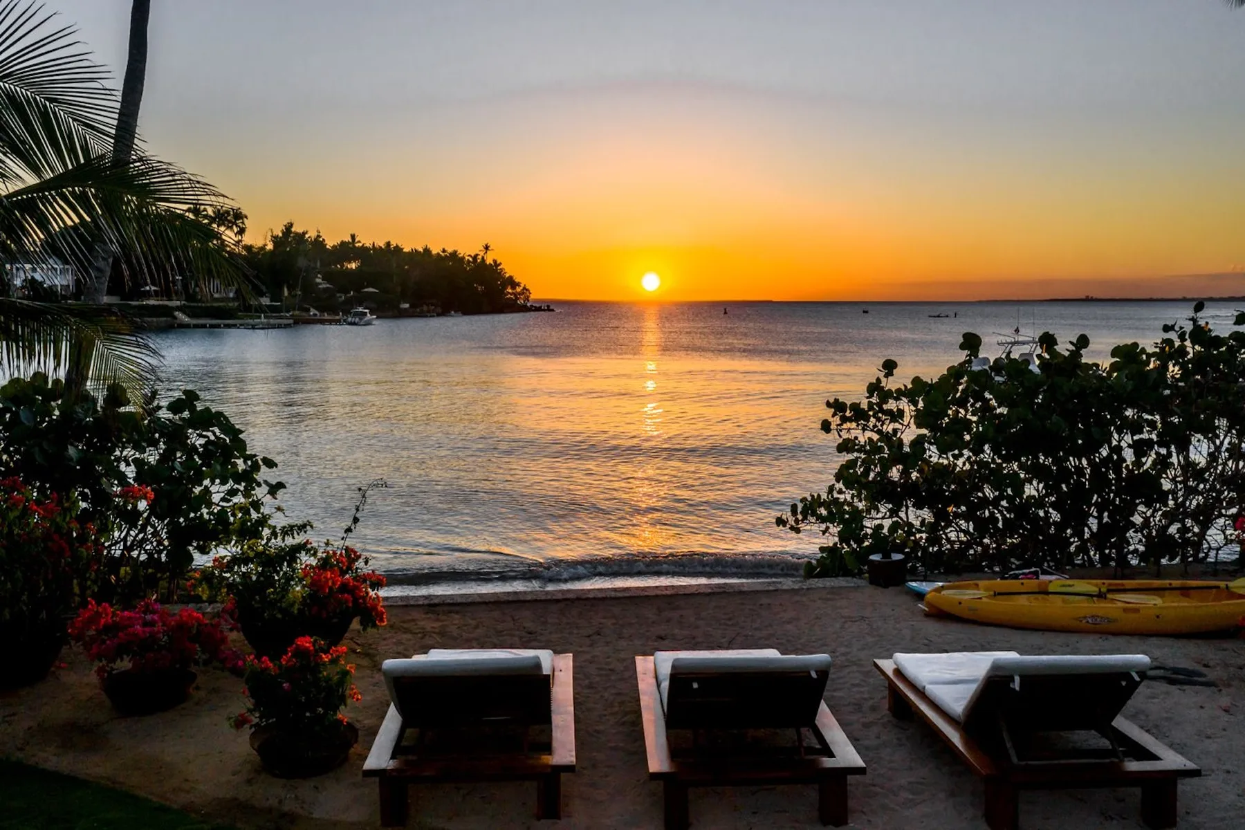 A sunset over calm water, viewed from a beach with lounge seating and flowering plants.