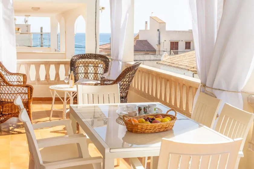 A sunlit balcony dining area with a table, seating, and views of the sea.
