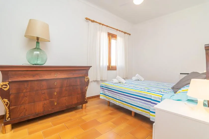 A bright bedroom with a striped bed beside a wooden chest of drawers.