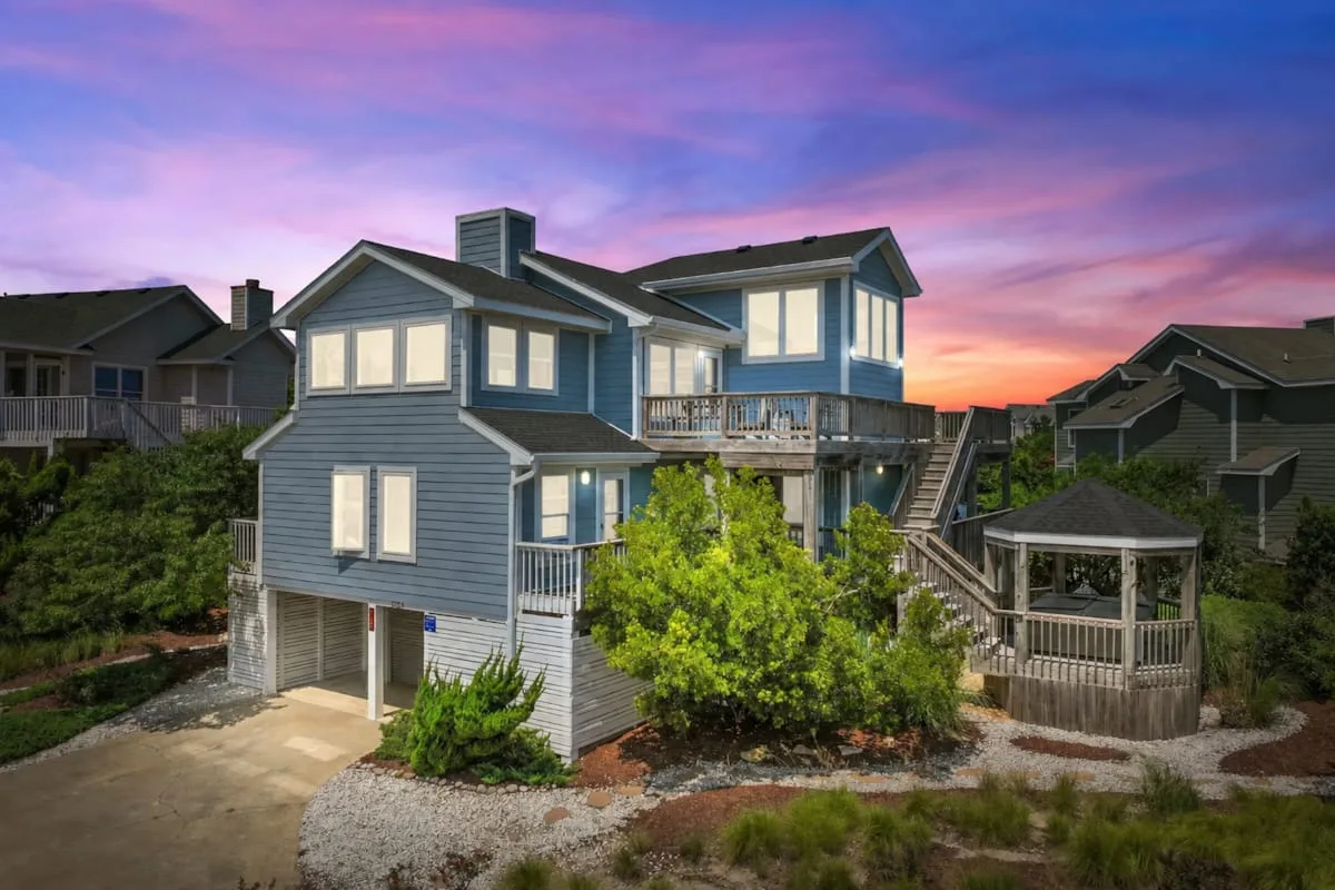 A large blue-gray house with wooden decks stands beneath a colorful sunset sky.