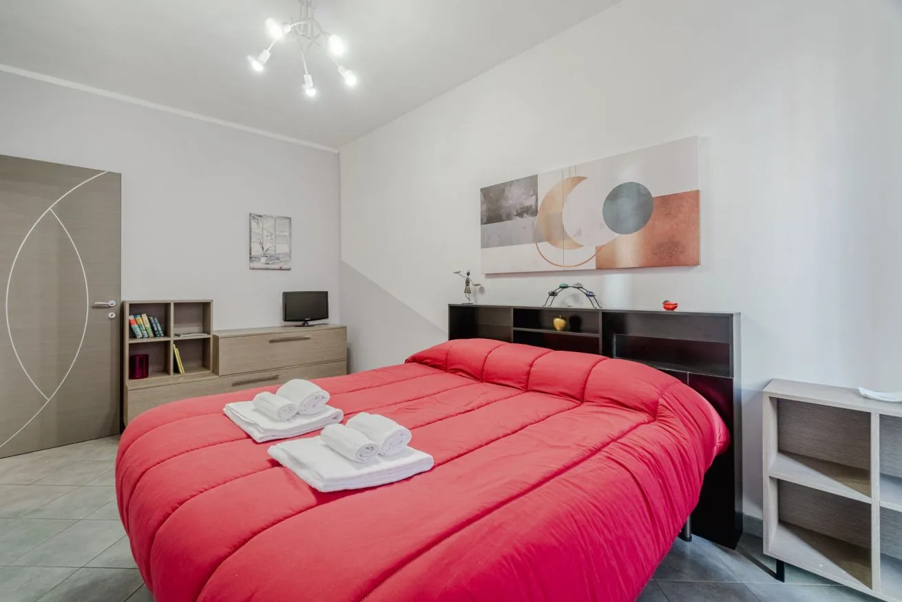 Un dormitorio con una cama roja, toallas dobladas y una obra abstracta en la pared.