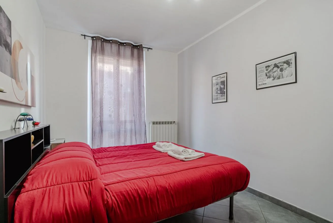 Un dormitorio con una cama roja, una ventana con cortinas y cuadros enmarcados.