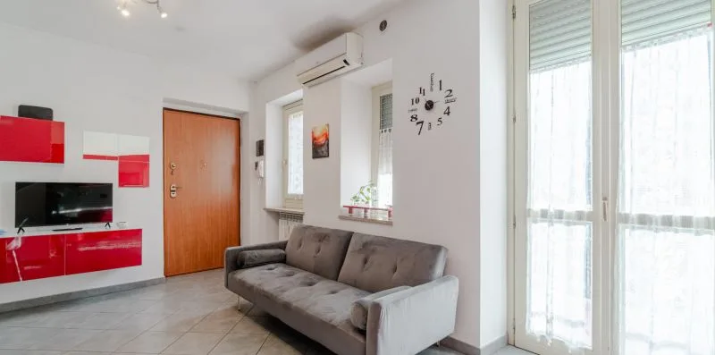 Una sala de estar con un sofá gris, un televisor en la pared y ventanas grandes.