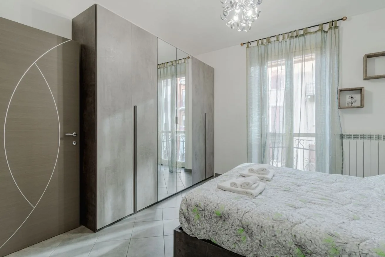 Un dormitorio luminoso con una cama grande junto a armarios con espejo y ventanas altas con cortinas.