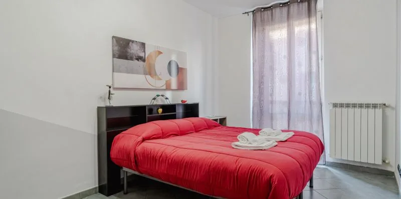 Un dormitorio con una cama roja y una cortina translúcida junto a la ventana.