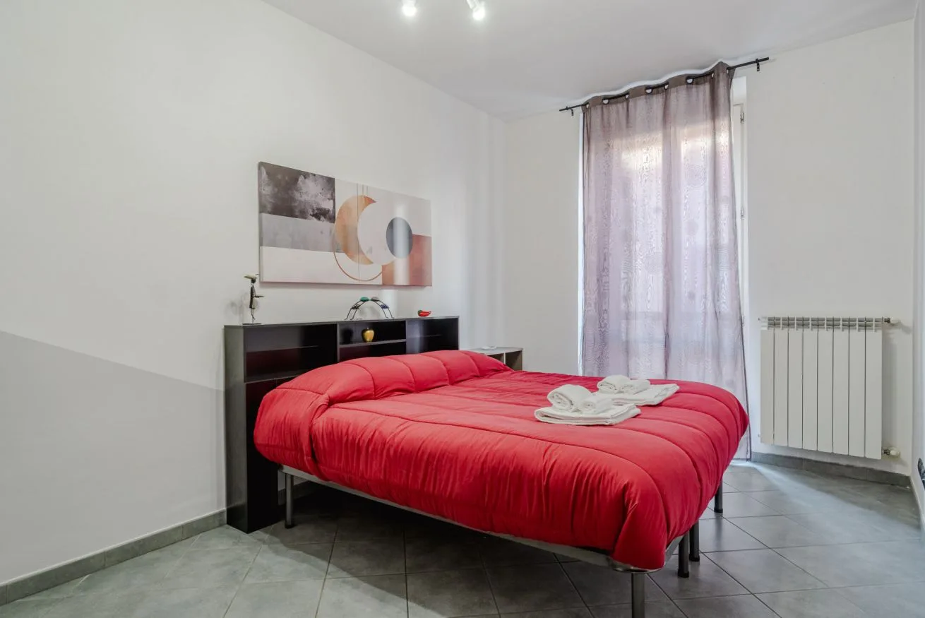 Un dormitorio con una cama roja, arte en la pared y una ventana con cortinas.