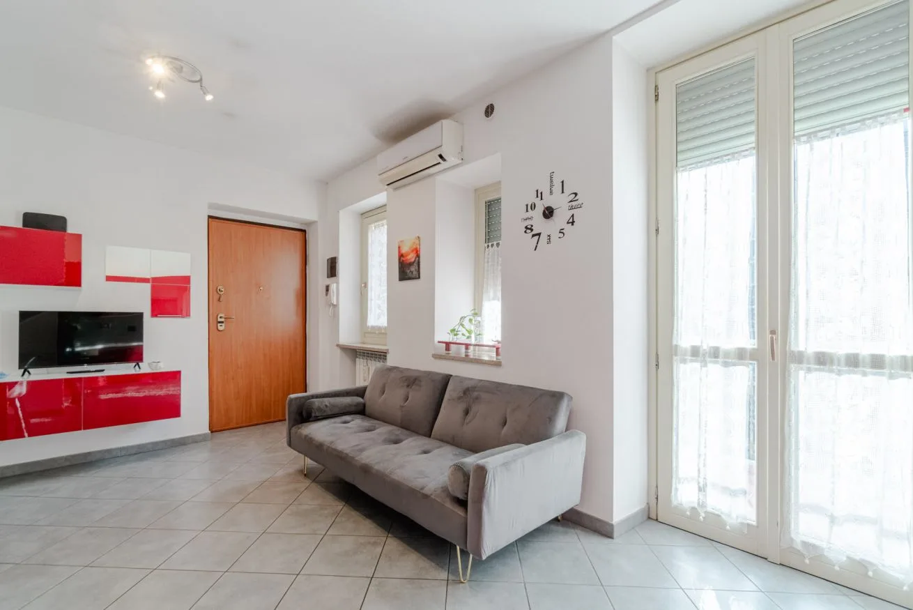 Una sala luminosa con un sofá gris, muebles rojos y ventanales grandes.