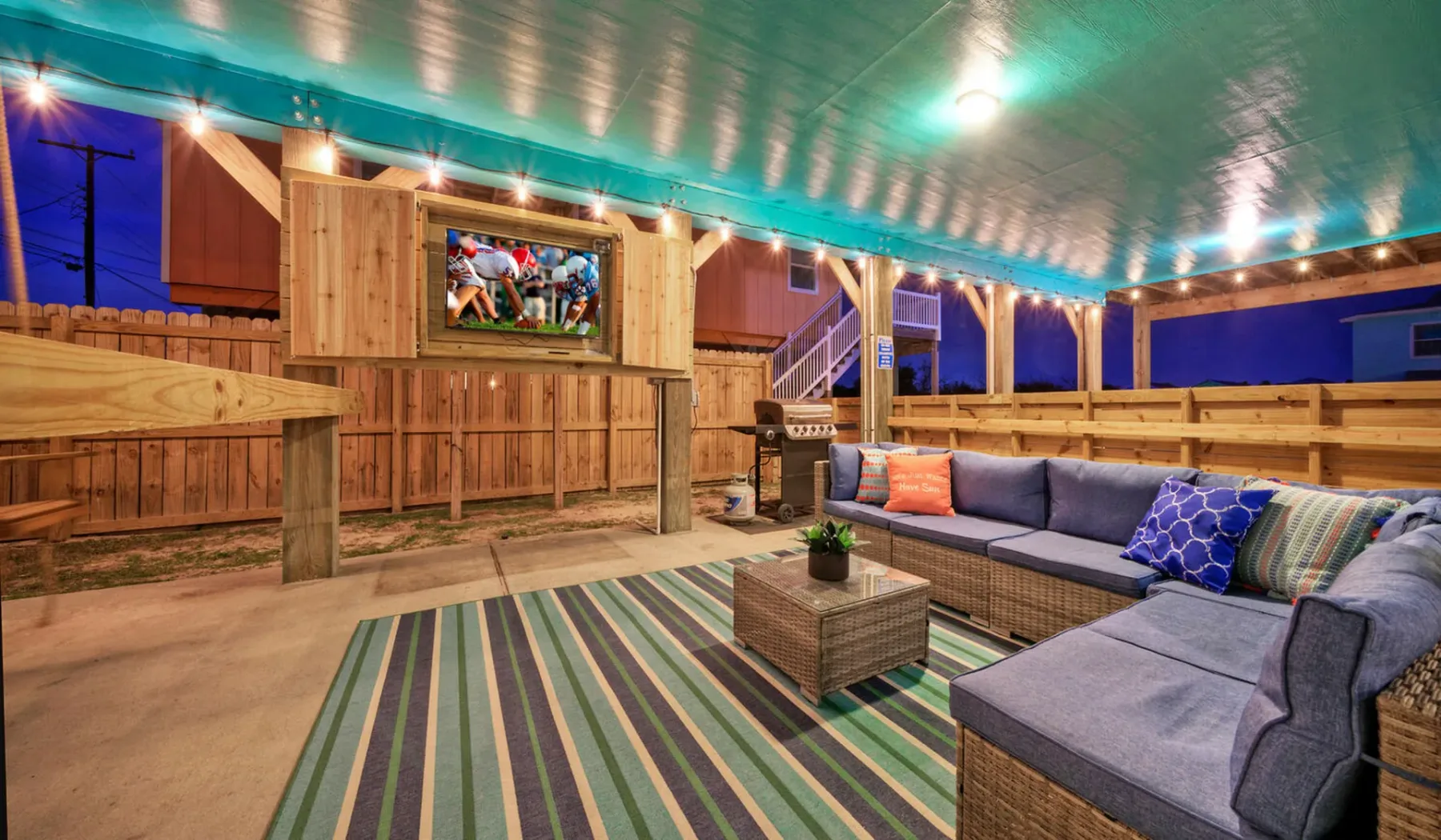 A covered outdoor seating area has a sectional sofa, coffee table, and striped rug.