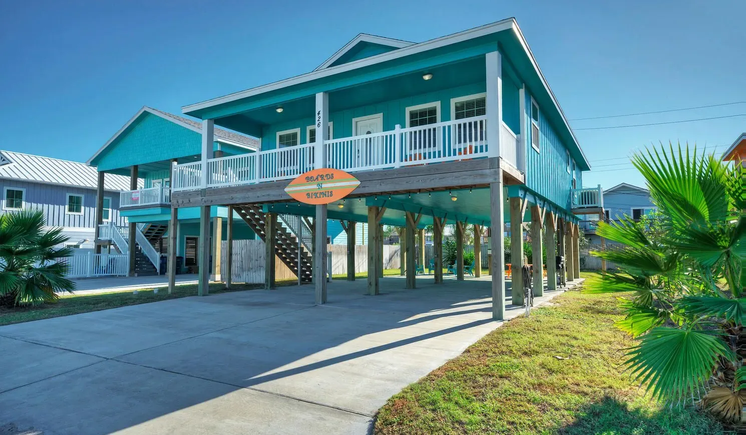 A raised turquoise coastal-style house with a wraparound balcony and open space beneath.