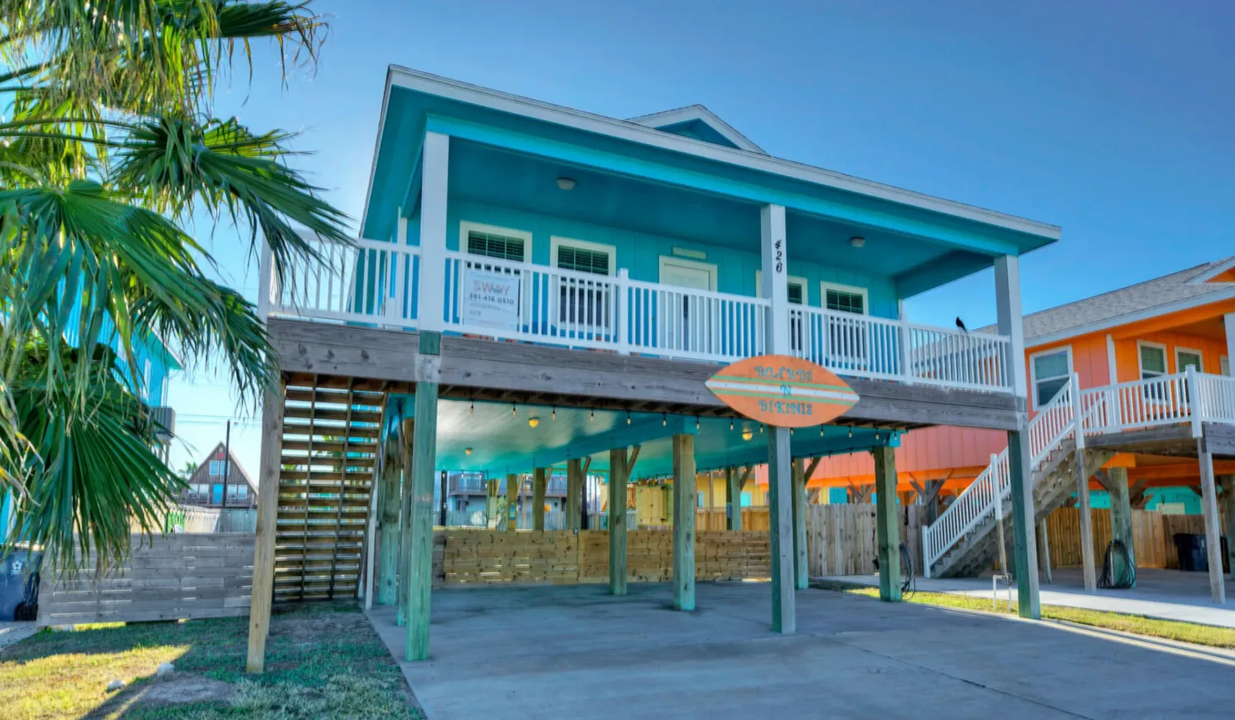 A raised turquoise building with a wraparound balcony stands beside a neighboring orange building.