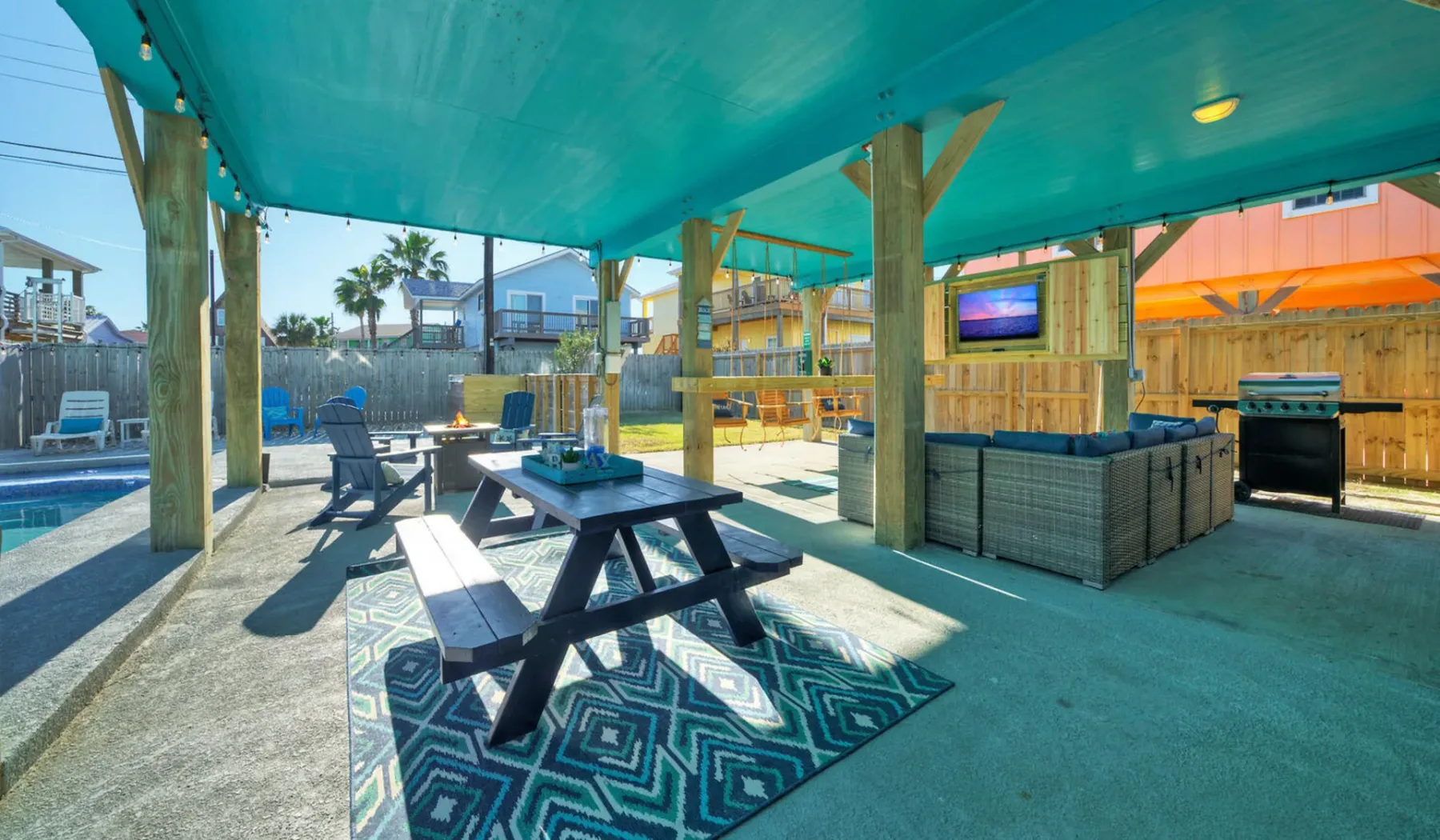 A covered outdoor patio has picnic seating, a television, and a pool nearby.