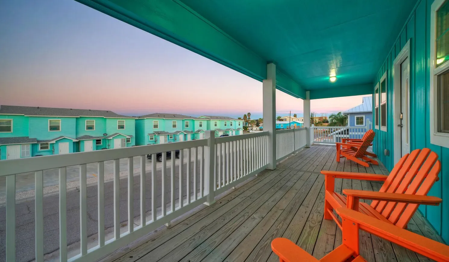 A covered wooden balcony with orange seating overlooks neighboring buildings at sunset.