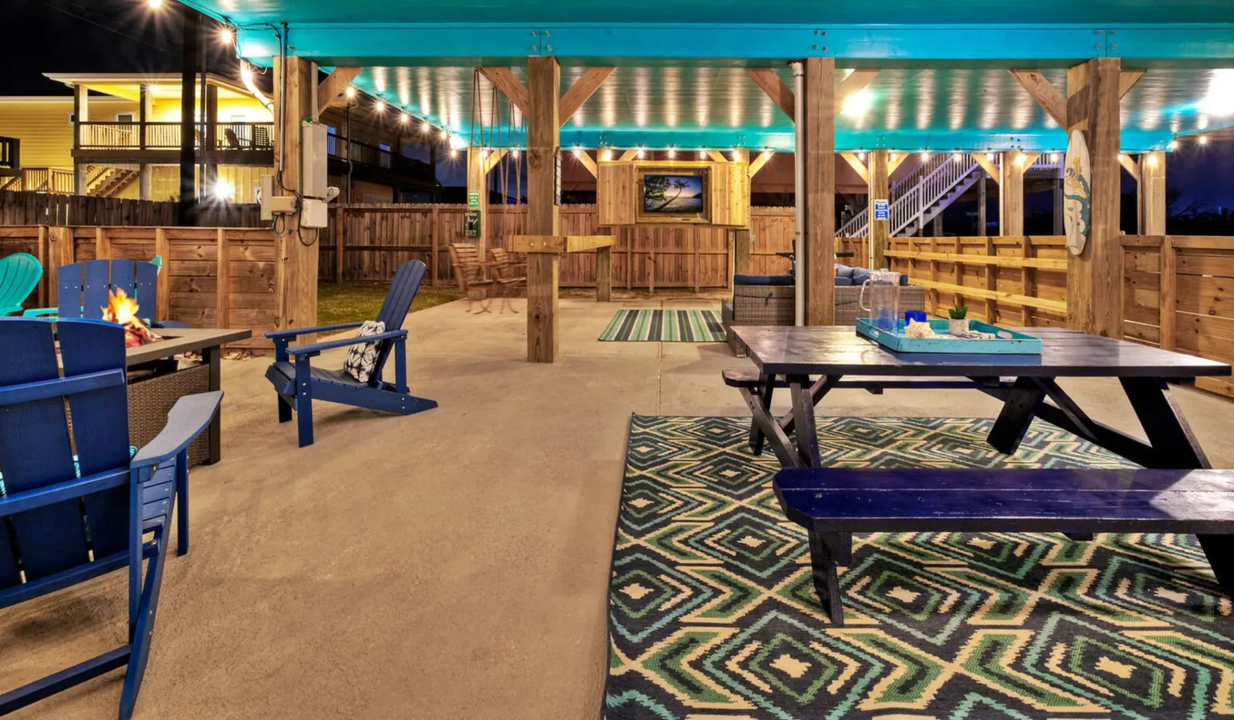 A covered outdoor seating area has picnic tables, chairs, and a patterned rug.
