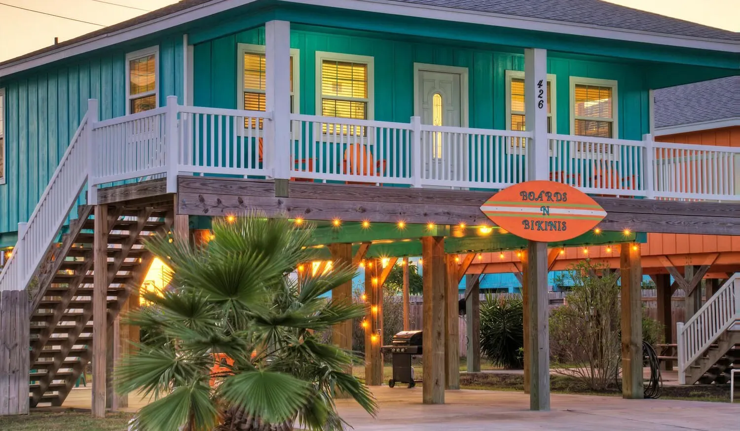 A raised teal building has a wooden balcony, exterior stairs, and warm lights.
