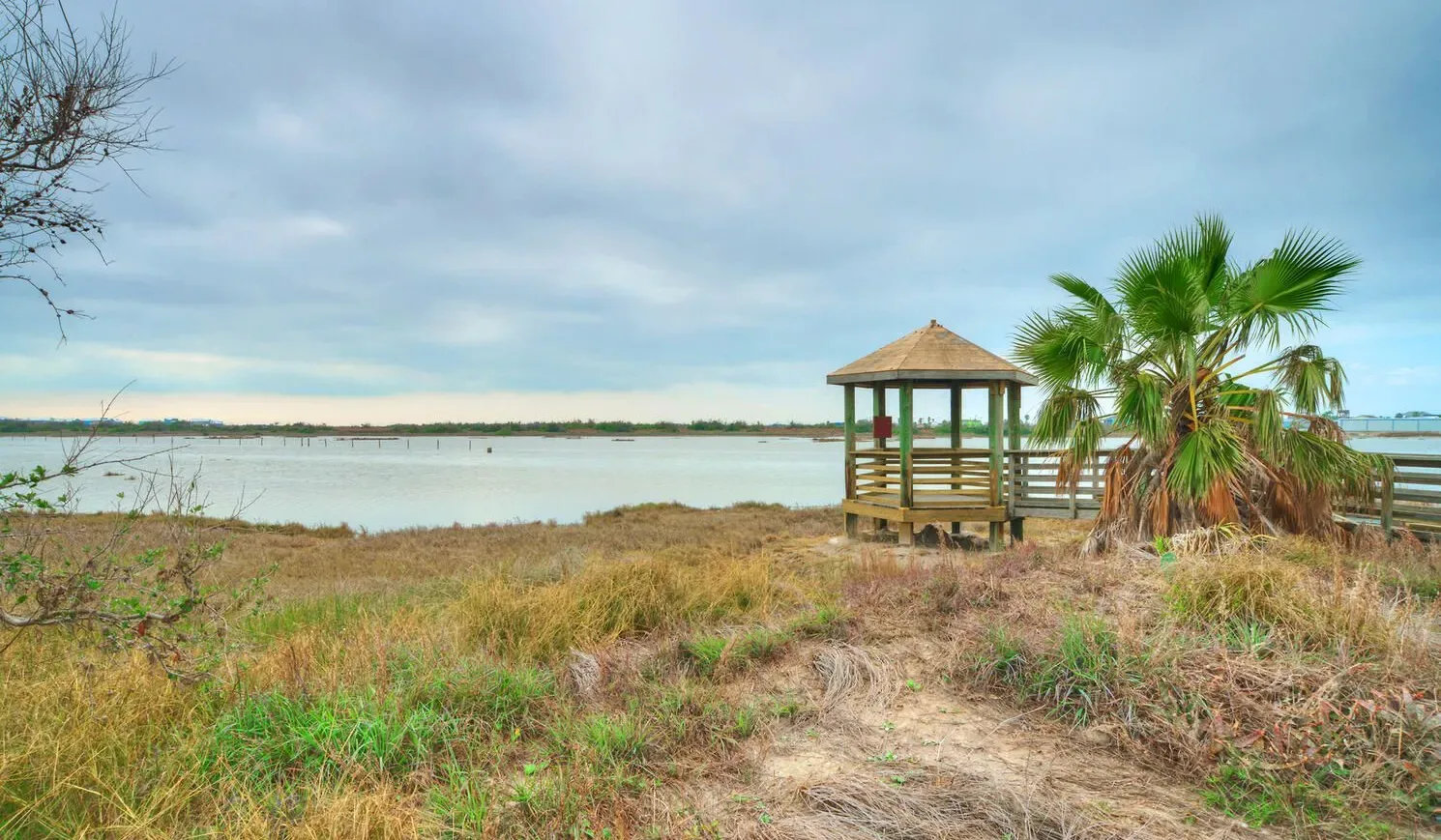 A wooden gazebo stands beside a calm body of water amid grassy vegetation.