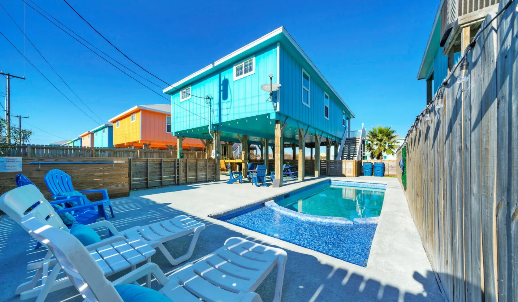 An outdoor pool sits beside a raised turquoise house, with lounge seating nearby.