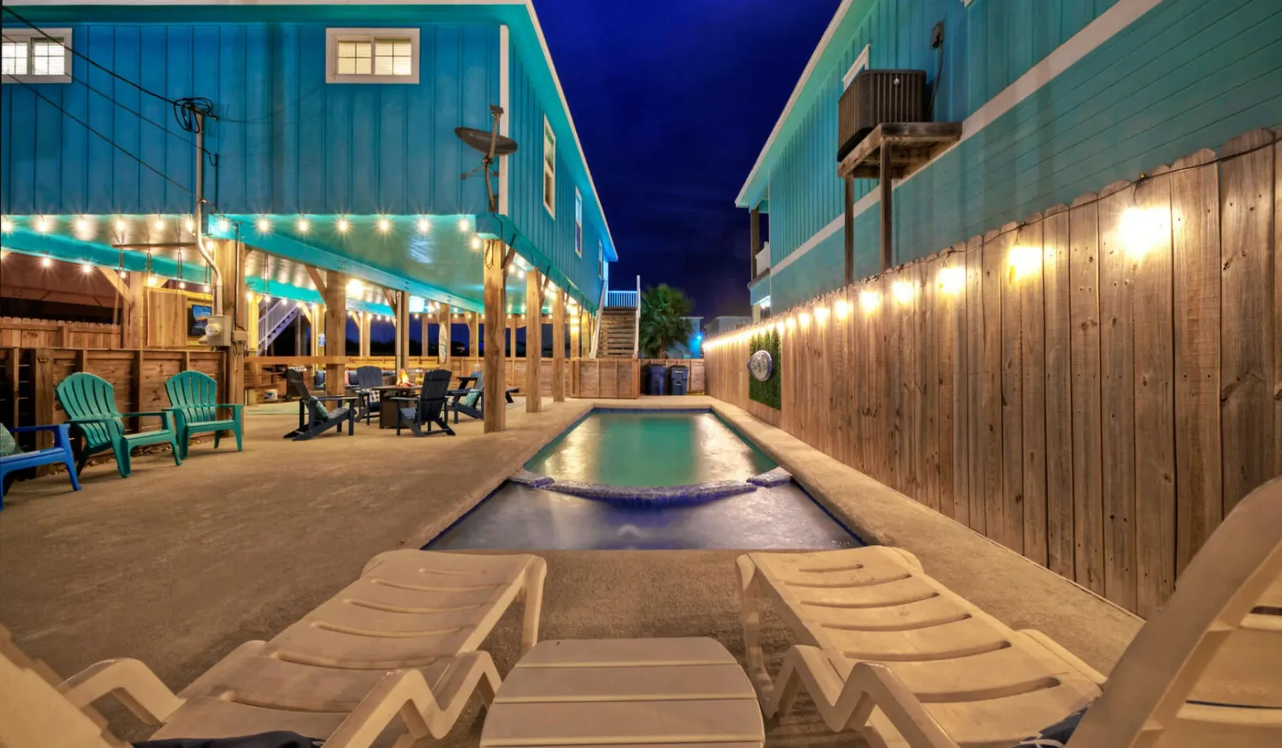 A lit outdoor pool sits between teal buildings, with lounge chairs in the foreground.