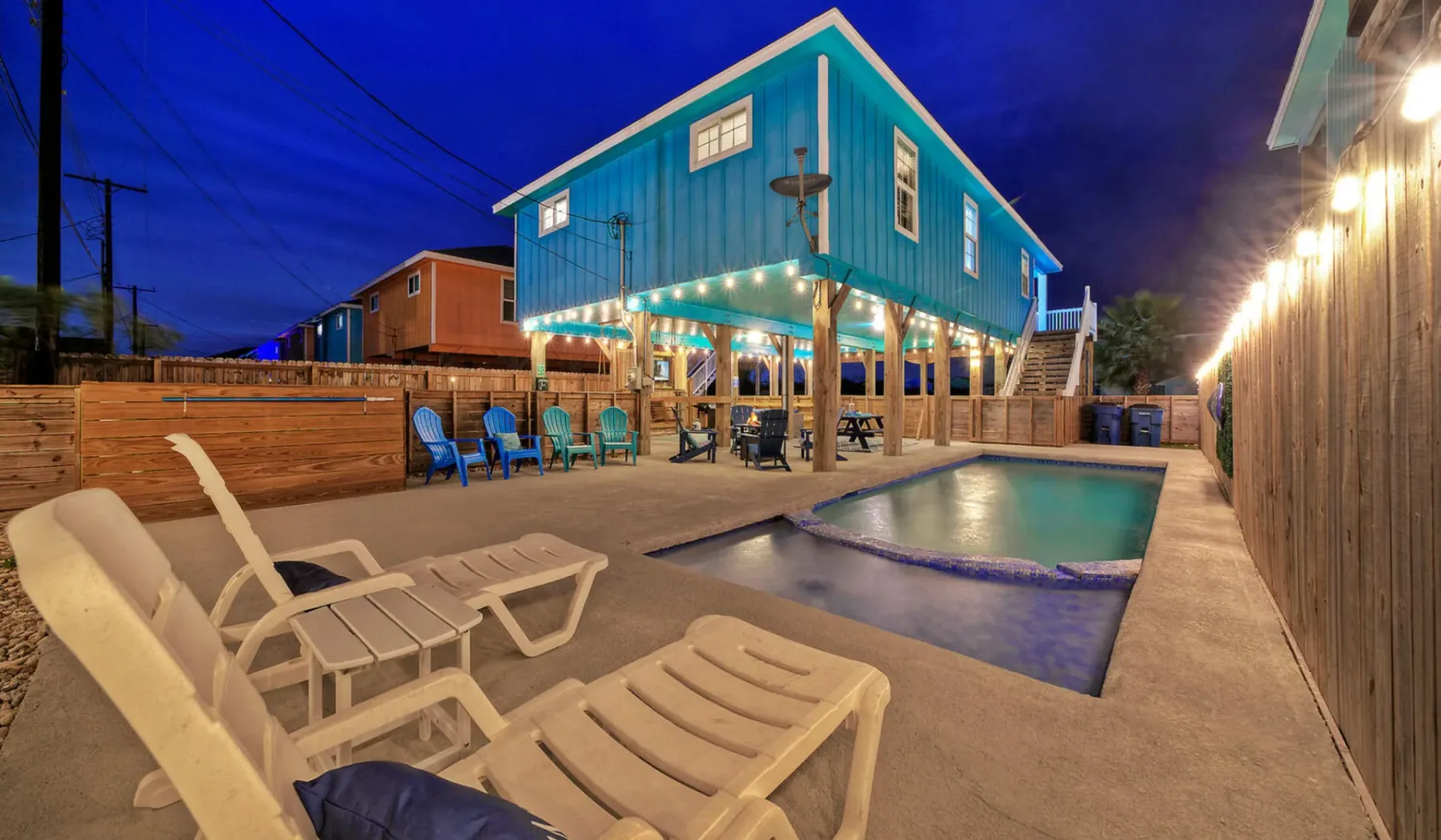 A nighttime pool patio with lounge seating beside an elevated blue building.