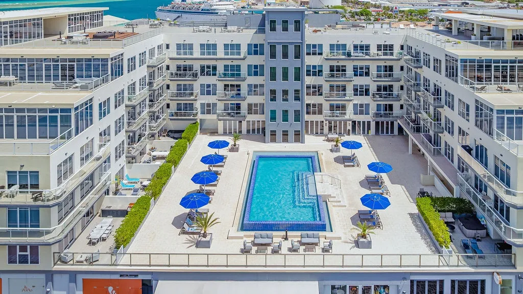 Aerial view of a modern property featuring rooftop pool deck with lounge chairs, umbrellas, and greenery surrounded by multi-story vacation rental buildings near the ocean.