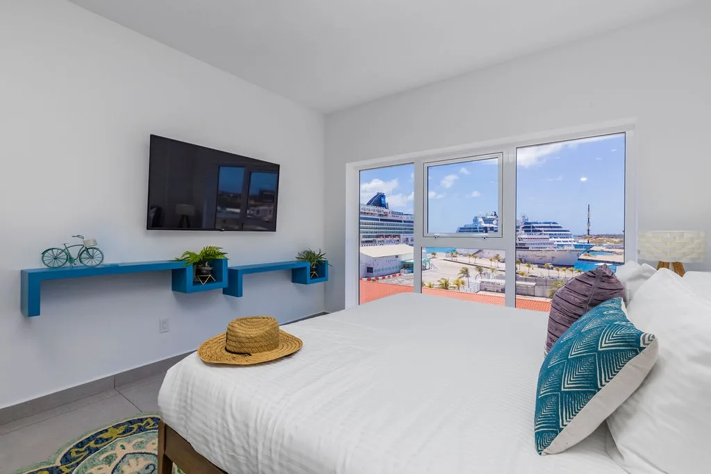 Crisp white linens and bright blue accents frame stunning harbor views through floor-to-ceiling windows. Relax and watch cruise ships glide by from your comfortable bed.