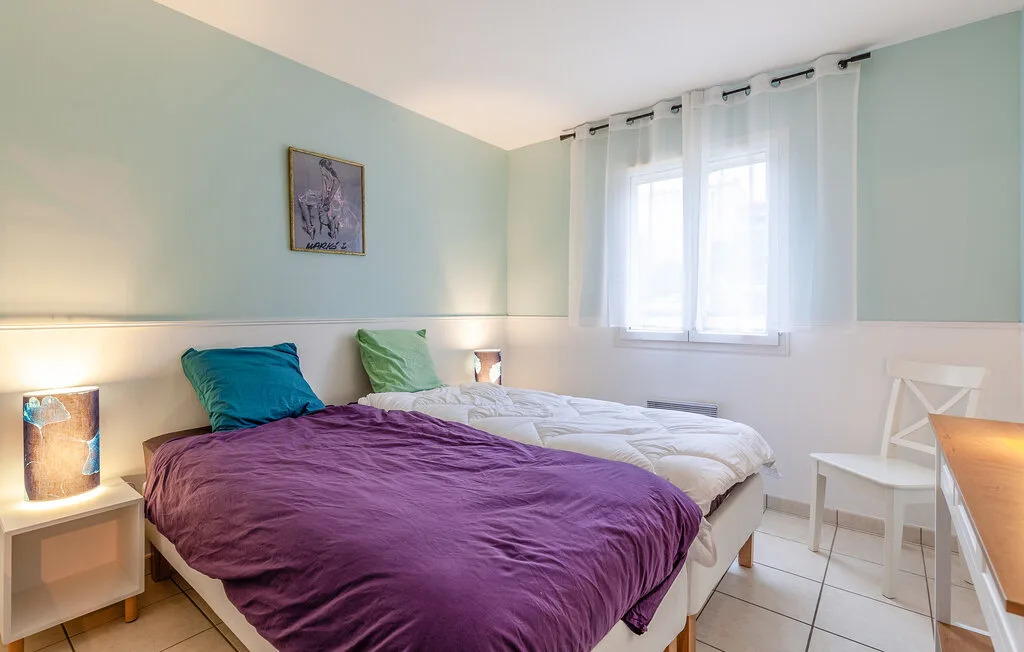 Bedroom with beds covered in purple and white bedding beside a curtained window.