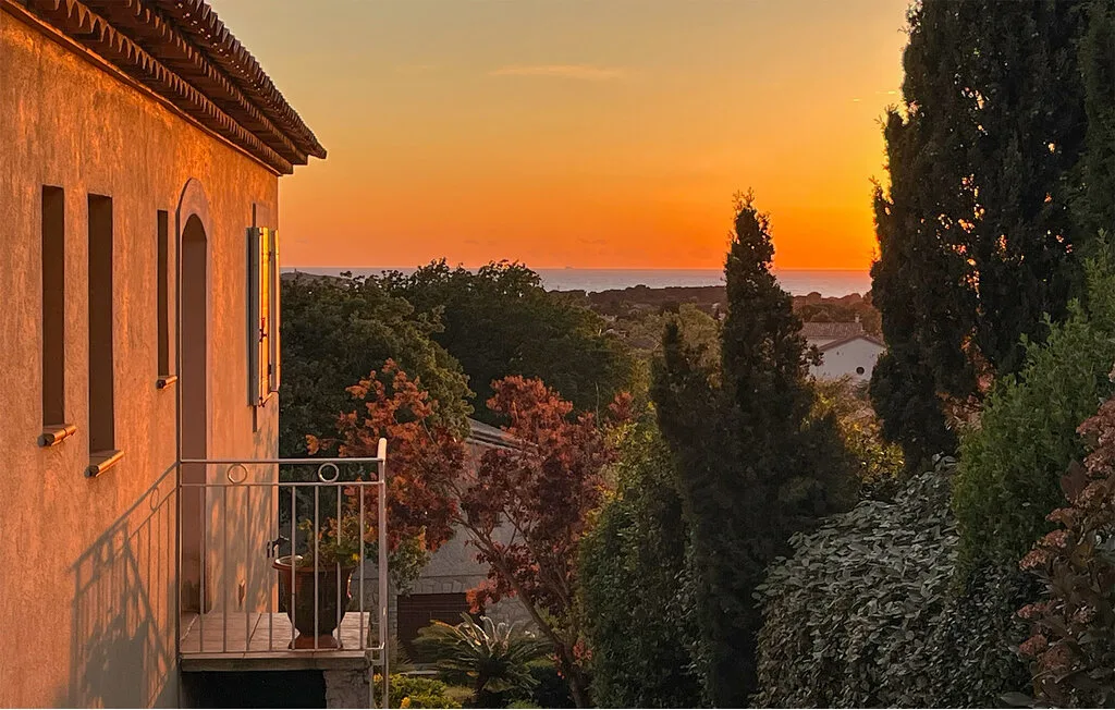 A sunset view from a balcony overlooks trees and distant rooftops.