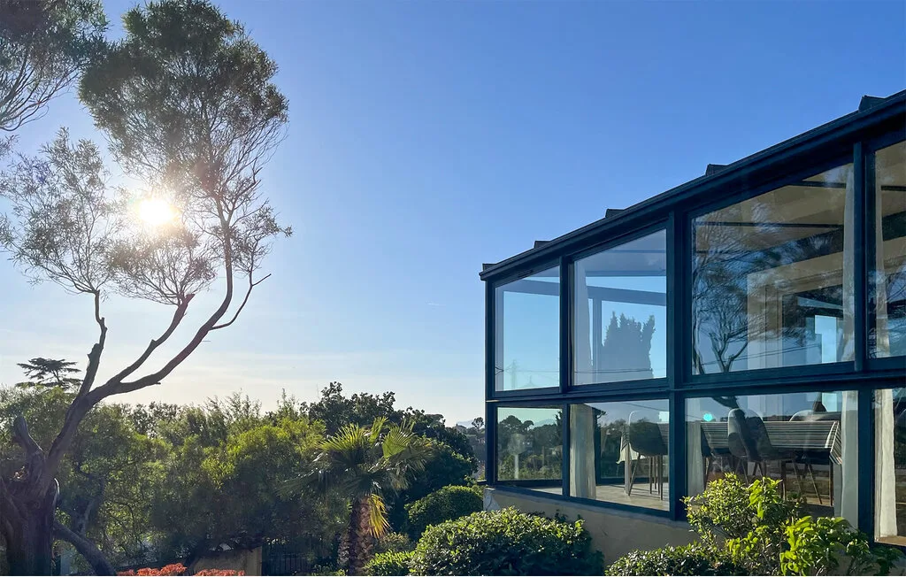 A glass-walled building stands beside leafy shrubs under a clear sky.