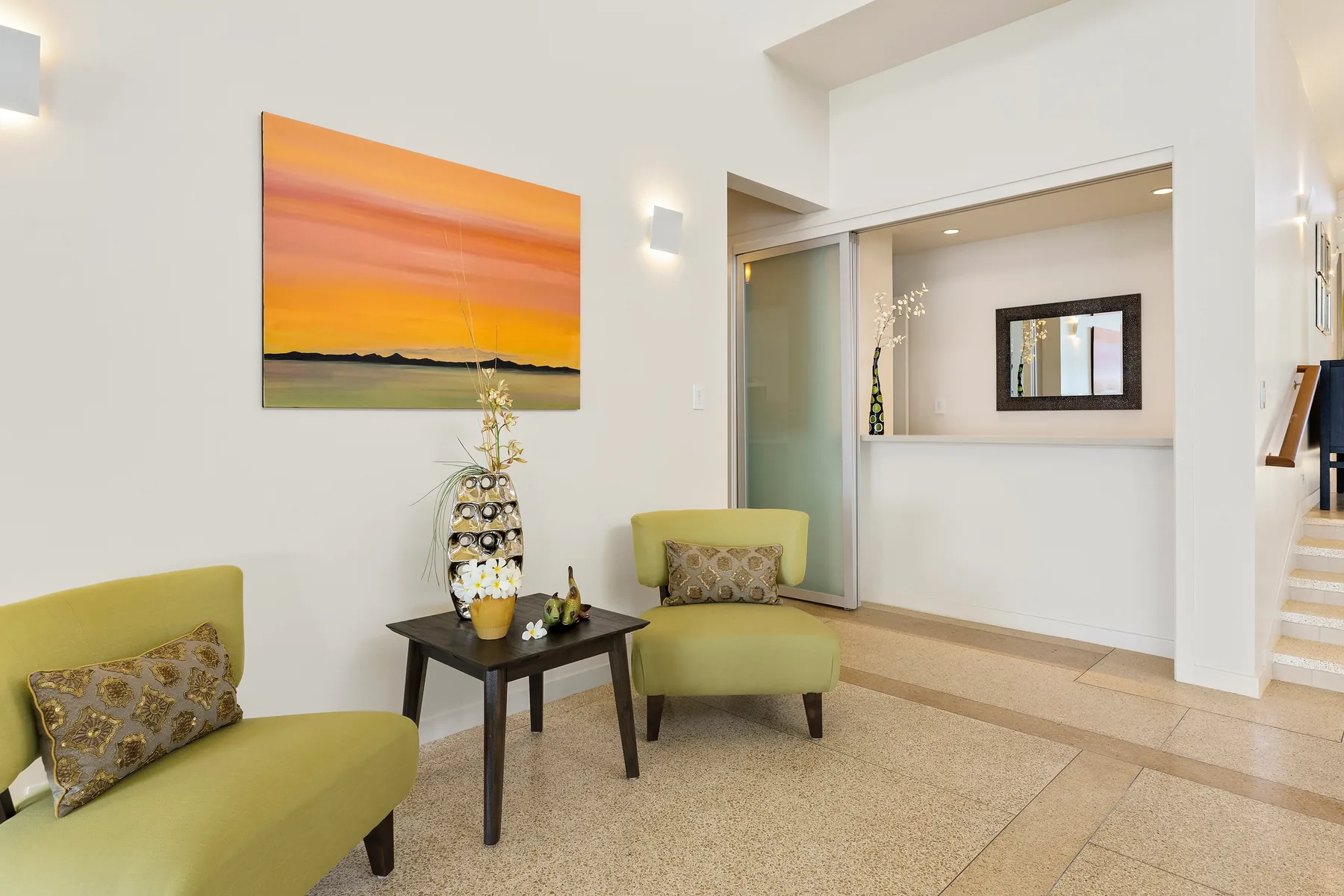 Una luminosa sala de estar con asientos verdes y una mesa pequeña bajo una pintura de atardecer.