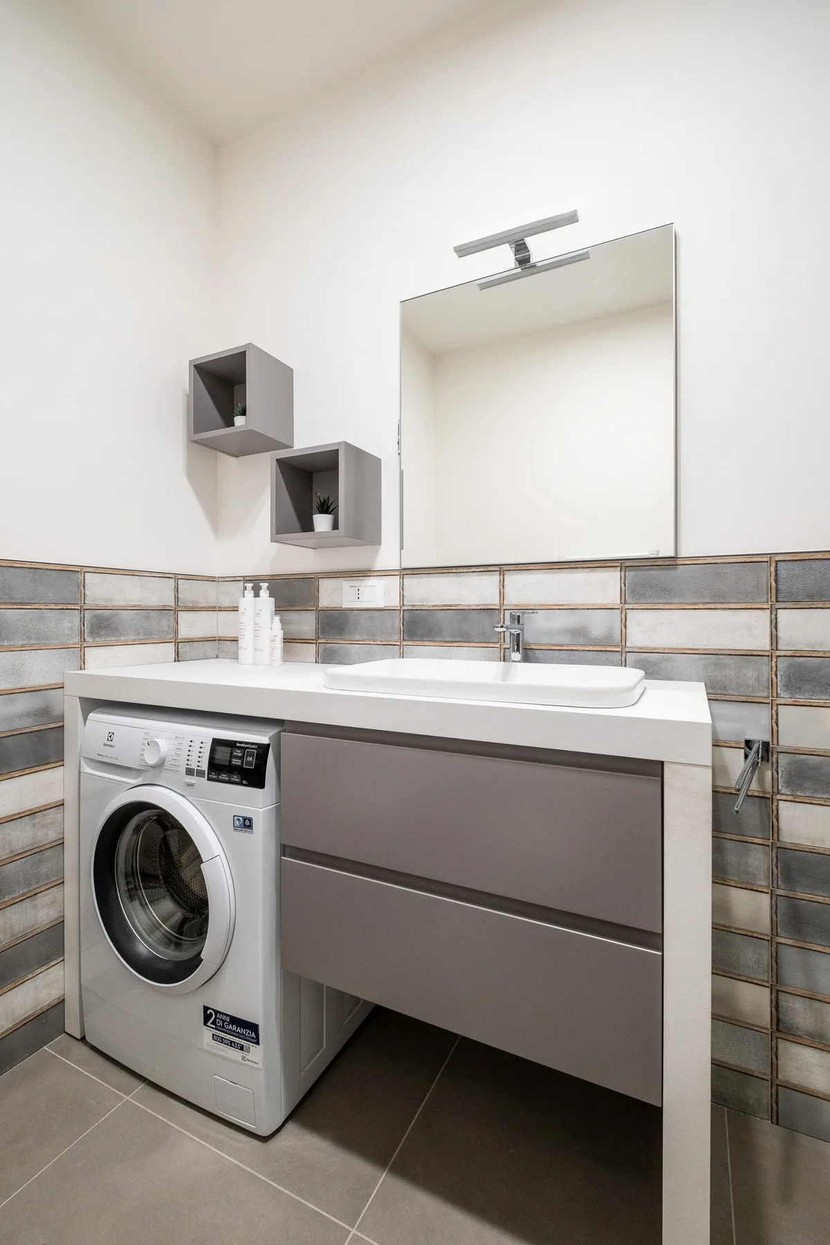 Una pequeña zona de lavandería con una lavadora de carga frontal bajo un lavabo y un espejo.