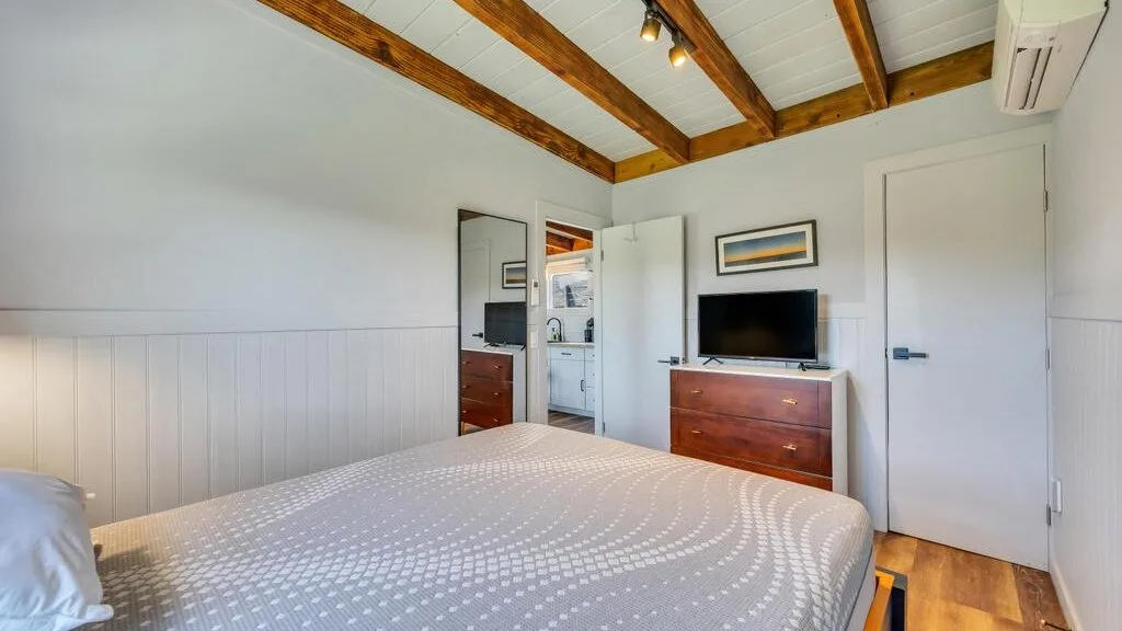 A bedroom with a bed, dresser, and television beneath exposed ceiling beams.