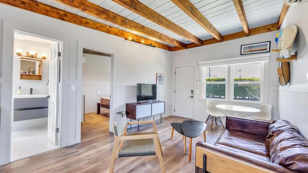A bright living room with exposed ceiling beams and seating beside the windows.