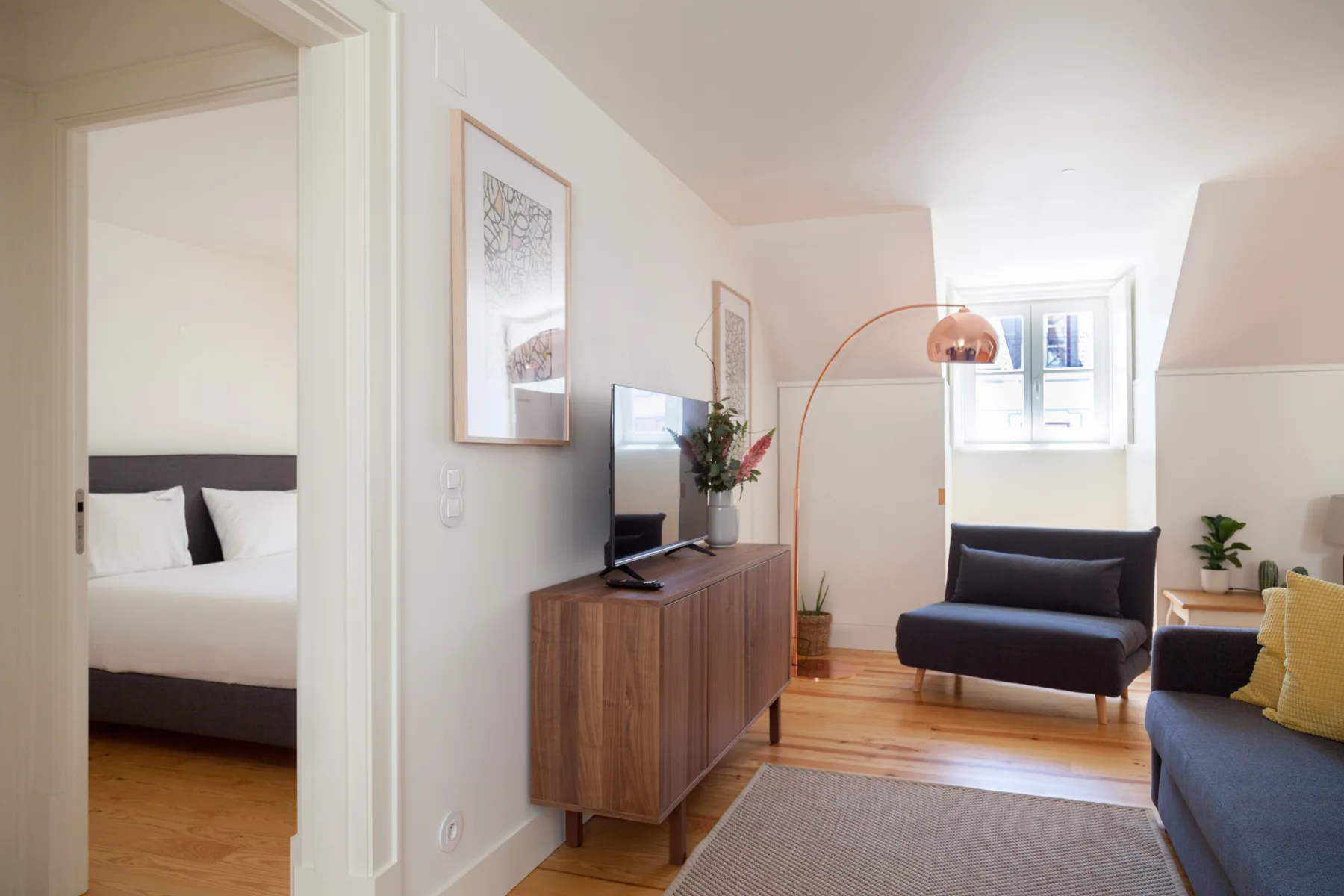 A bright living room with seating, a television, and a view into a bedroom.