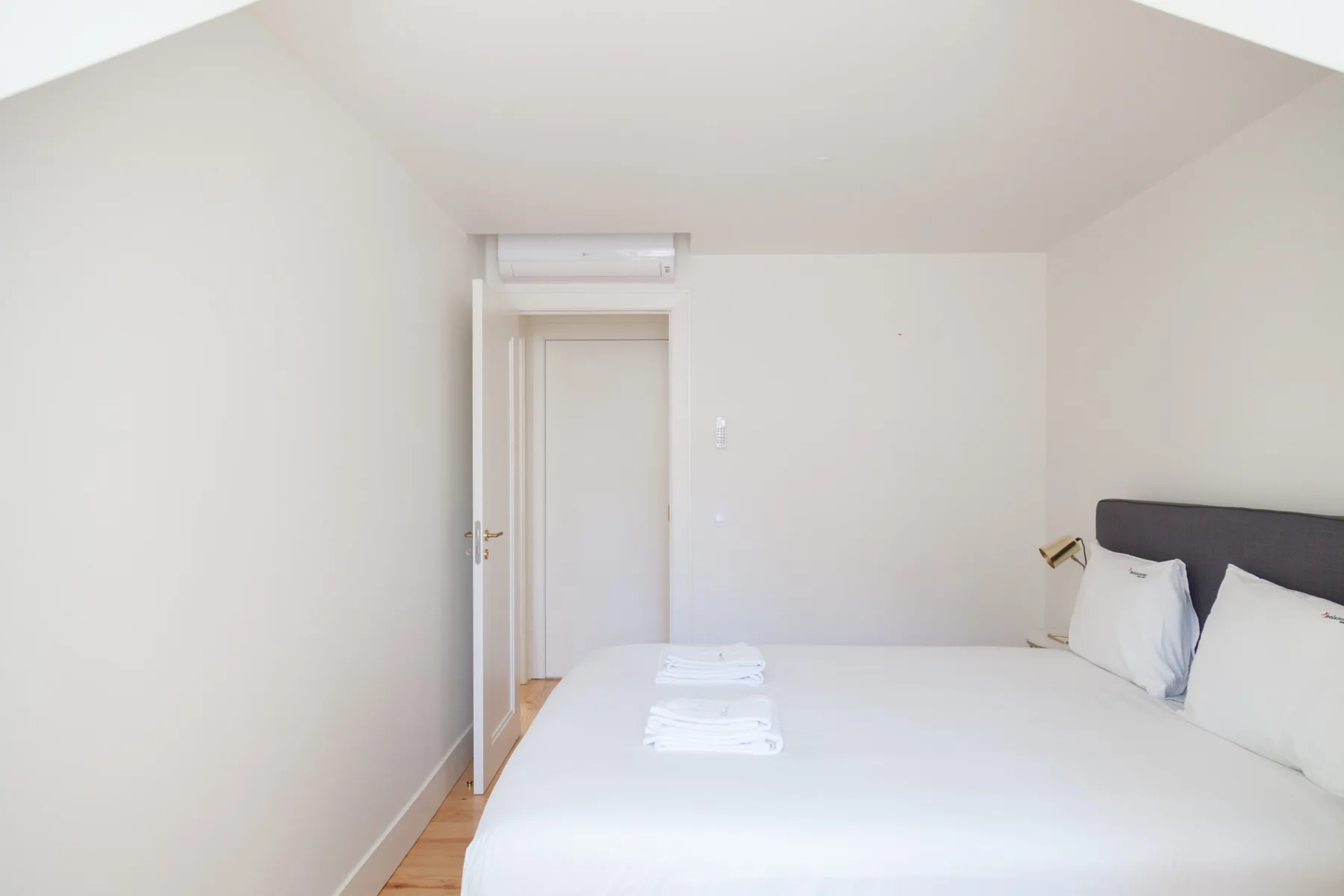 A bright bedroom with a bed, folded towels, and a wall-mounted air conditioner.