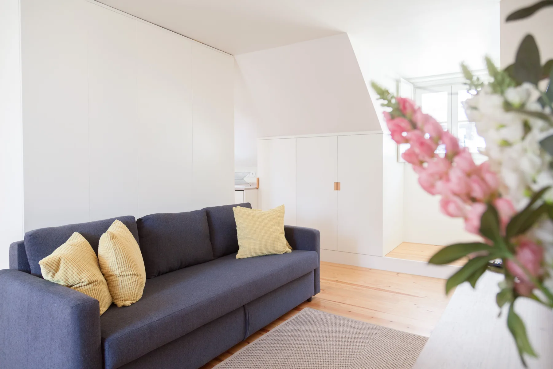 A bright living room with a dark sofa, pale cushions, and flowers in the foreground.