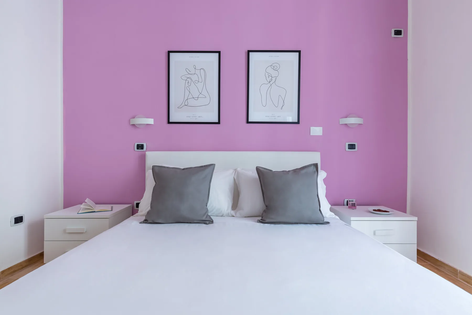 A bedroom with a neatly made bed against a pink wall with framed artwork.