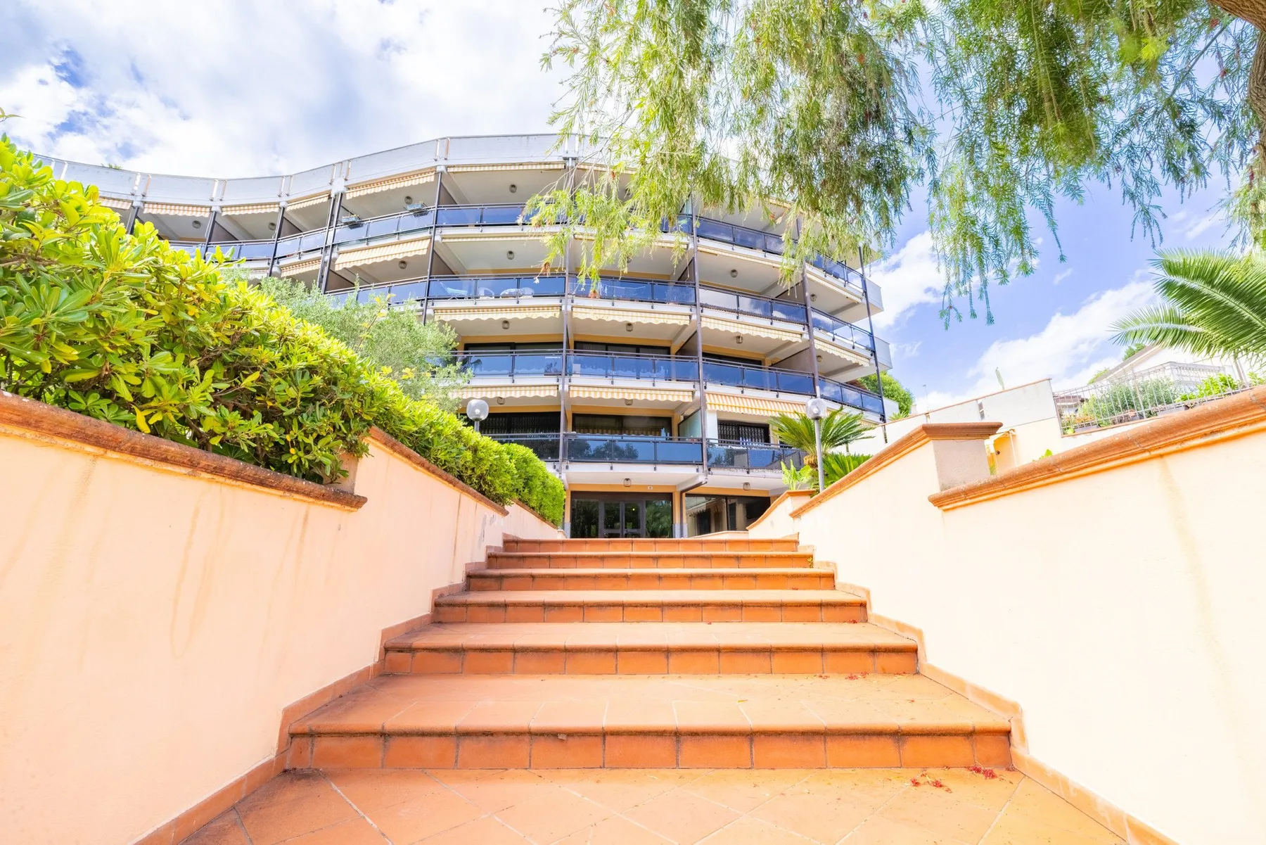 Escalones de terracota conducen hacia un edificio de varias plantas entre vegetación.
