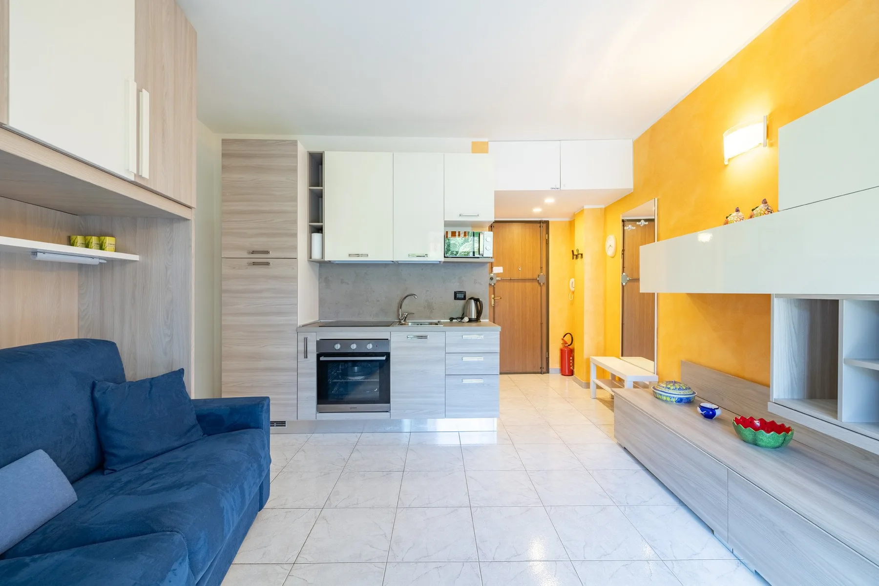 Una sala luminosa se abre a una cocina compacta, con asientos azules y gabinetes blancos.