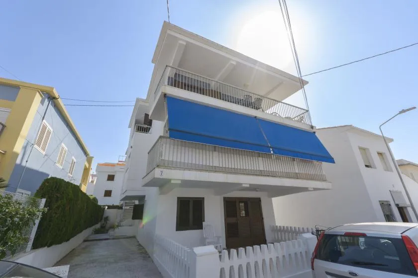 A white multi-level building with blue balcony awnings stands beside a narrow street.