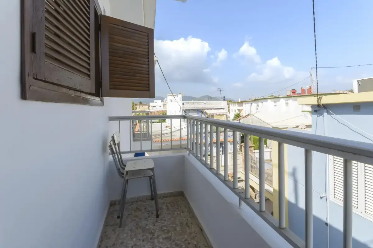 A narrow balcony has metal railings, a chair, and views of nearby buildings.