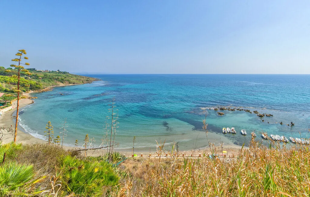 A coastal bay with clear turquoise water, rocky shoreline, and grasses in the foreground.