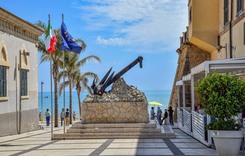 A seaside plaza features a large anchor monument, palm trees, and flags.