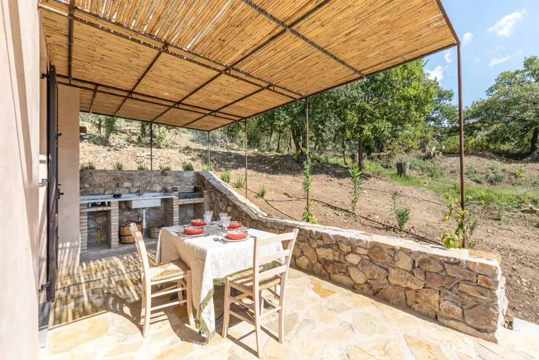 Una zona de comedor exterior a la sombra, con una mesa junto a una barbacoa de piedra.