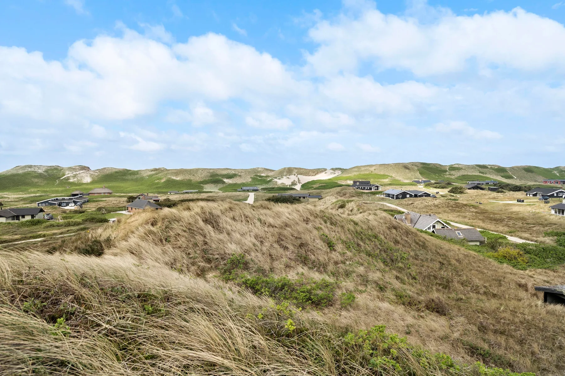 Un paisaje de dunas cubiertas de hierba con edificios dispersos bajo un cielo parcialmente nublado.