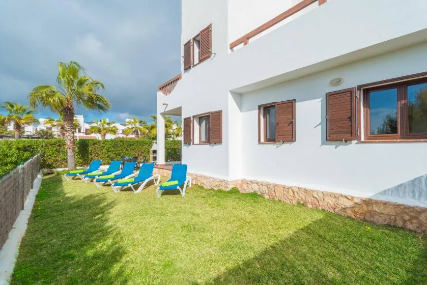 Una casa blanca junto a un jardín de césped, con asientos azules y palmeras.