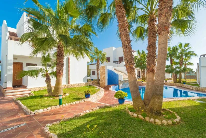A landscaped outdoor pool area with palm trees, greenery, and white buildings.