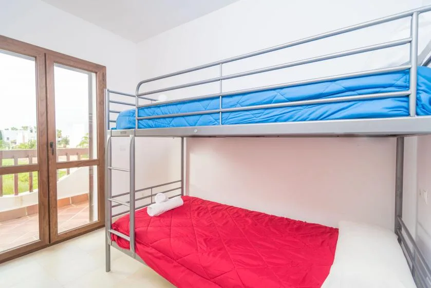 Un dormitorio con una litera metálica, ropa de cama azul arriba y roja abajo, junto a puertas de vidrio.