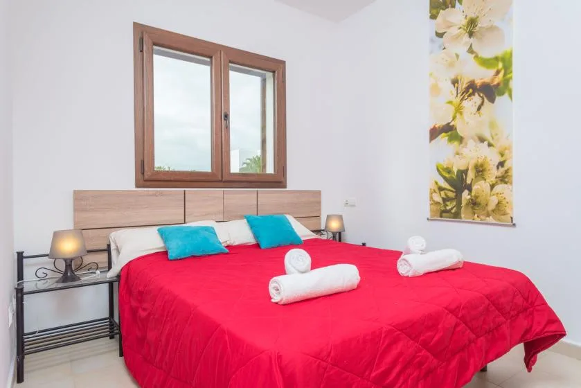 Un dormitorio con una colcha roja, almohadas azules y una ventana sobre el cabecero.